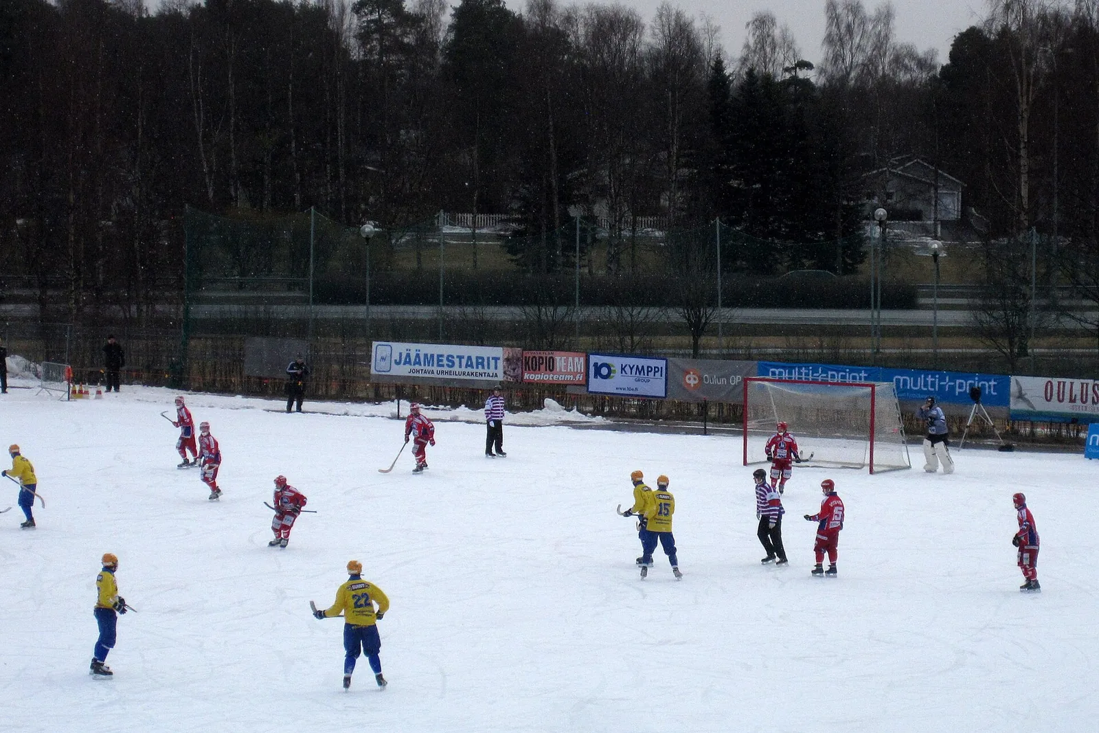 Jääpallo-ottelu ulkojäällä, pelaajat keltaisissa ja punaisissa asuissa