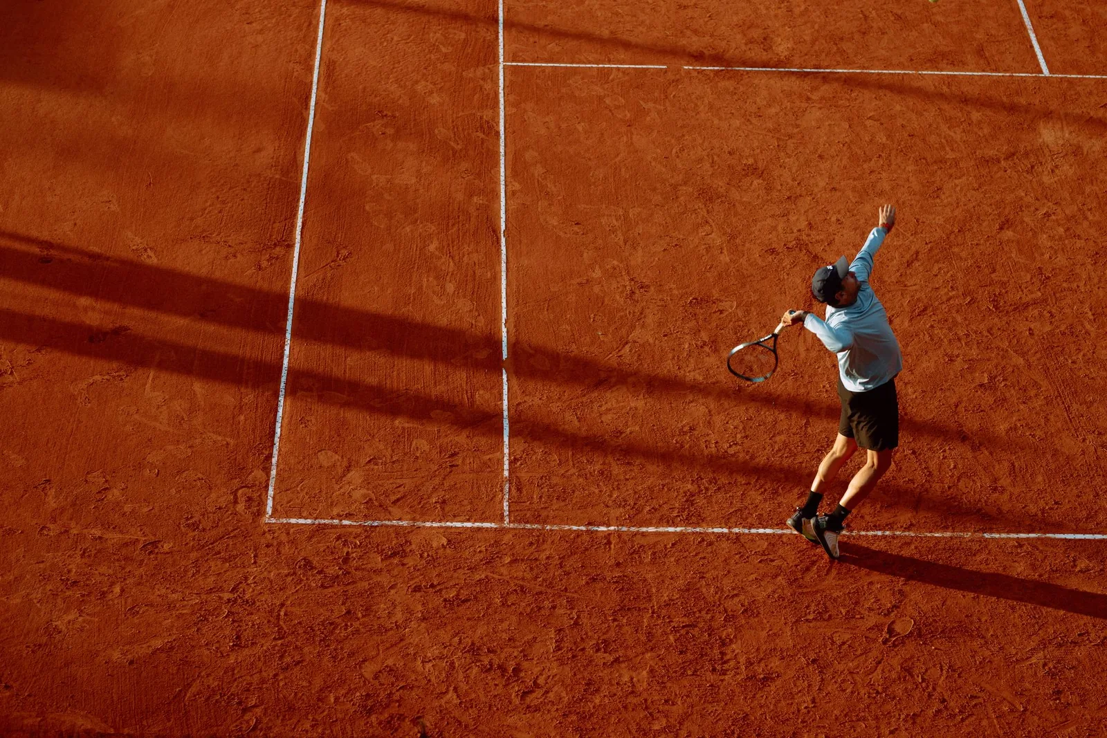 Tennispelaaja syöttää punasoraisella kentällä auringonlaskun valossa