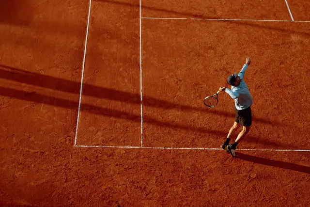 Tennispelaaja syöttää punasoraisella kentällä auringonlaskun valossa