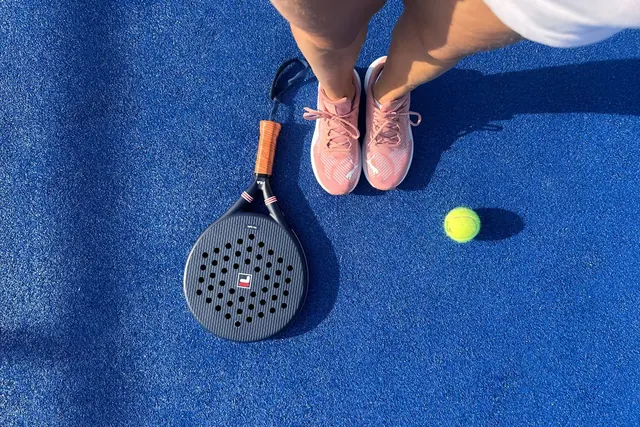 Vaaleanpunaiset urheilukengät, padelmaila ja pallo sinisellä kentällä