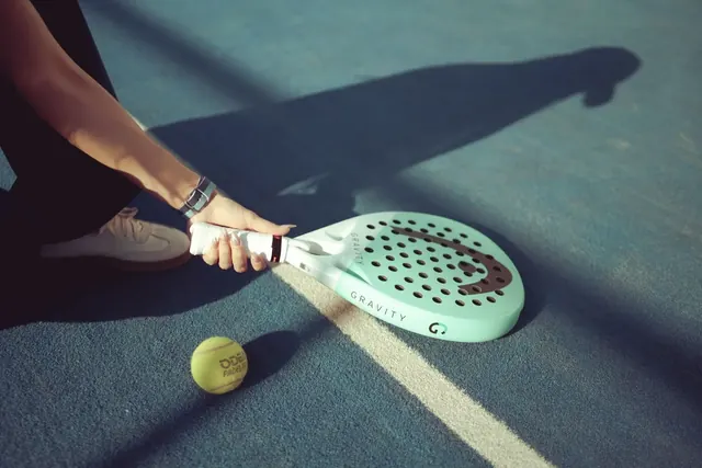 Käsi pitelee turkoosia padelmailaa kentällä, vieressä tennispallo